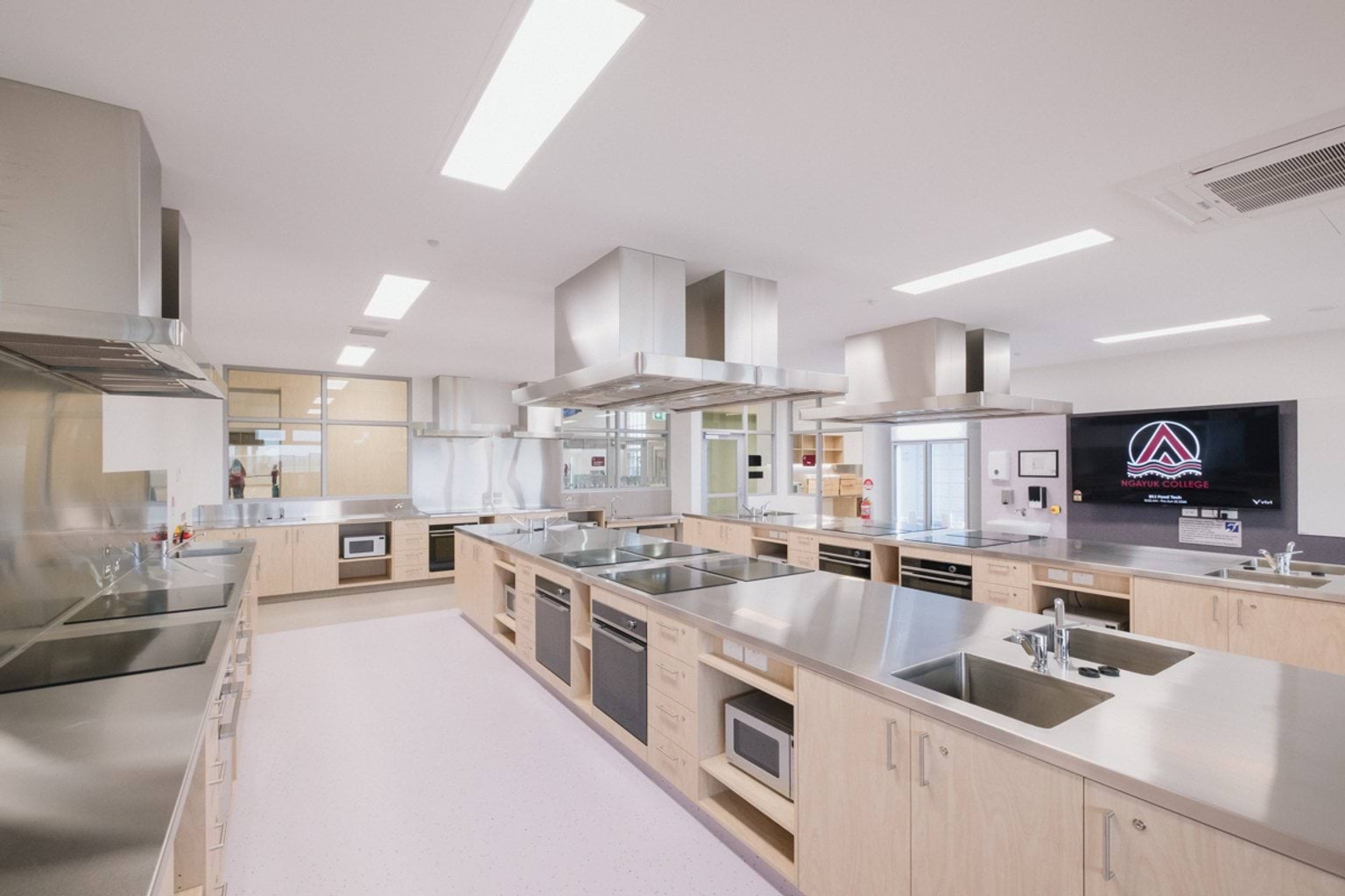 Food tech classroom with rows of metal counter tops each fitted with extraction fans, sinks, ovens, stoves and microwaves. We can see a screen on the far wall with the school logo and windows