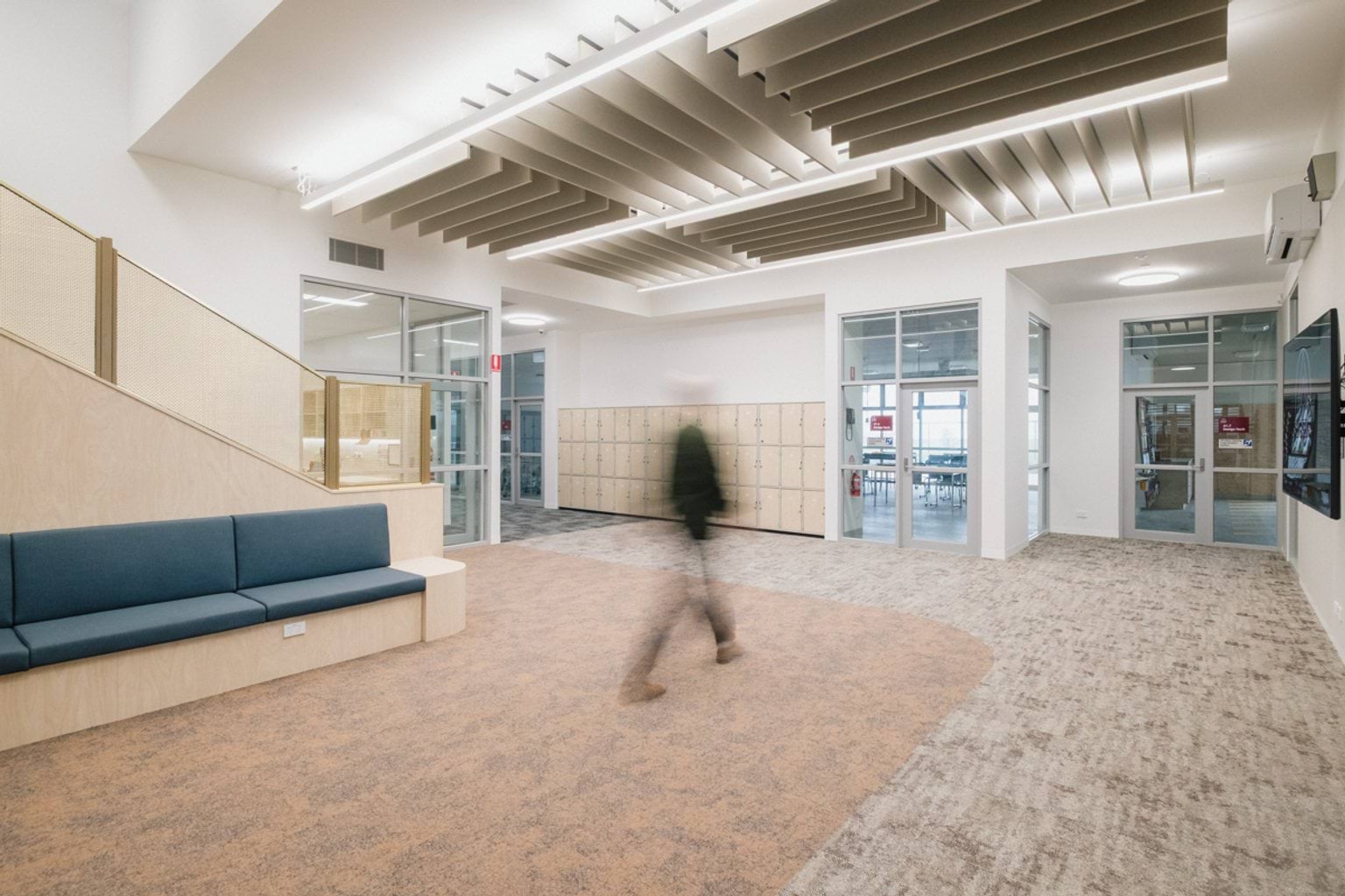 Open carpeted foyer area with a blurred person walking through. We can see a cushioned bench on the left with a stairwell behind, lockers across the far wall as well as hallways and doorways