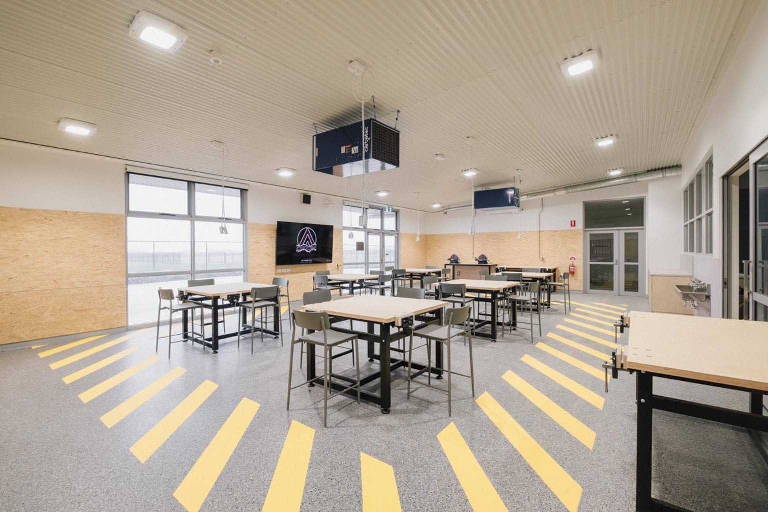 Workshop classroom with high tables and chairs, power cords hanging from the ceiling and yellow striped walkway surrounding the centre of the room