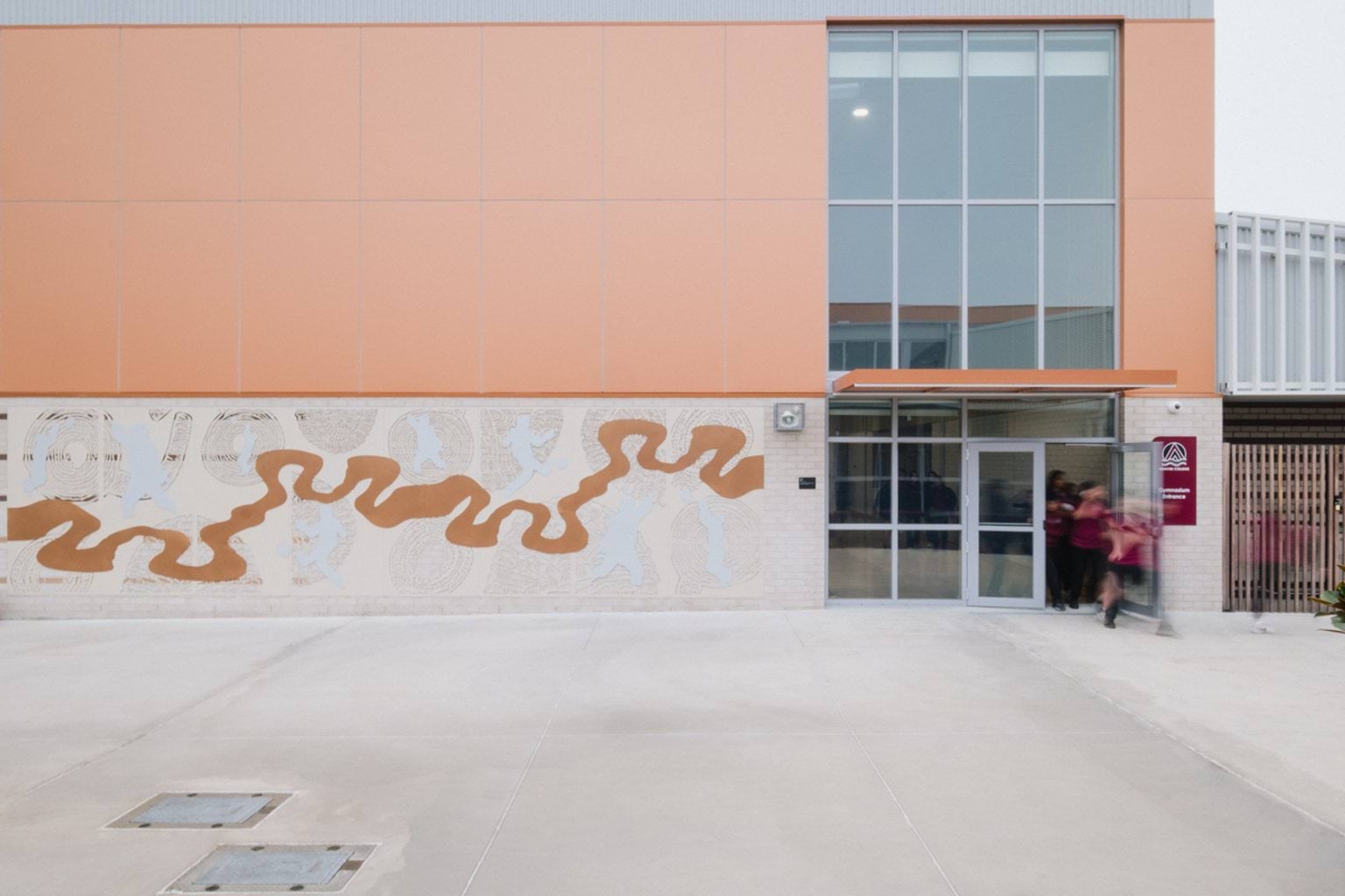 Outdoor paved area showing side entrance of building with abstract orange artwork on building wall and large windows. We can see blurred students exiting through the doorway