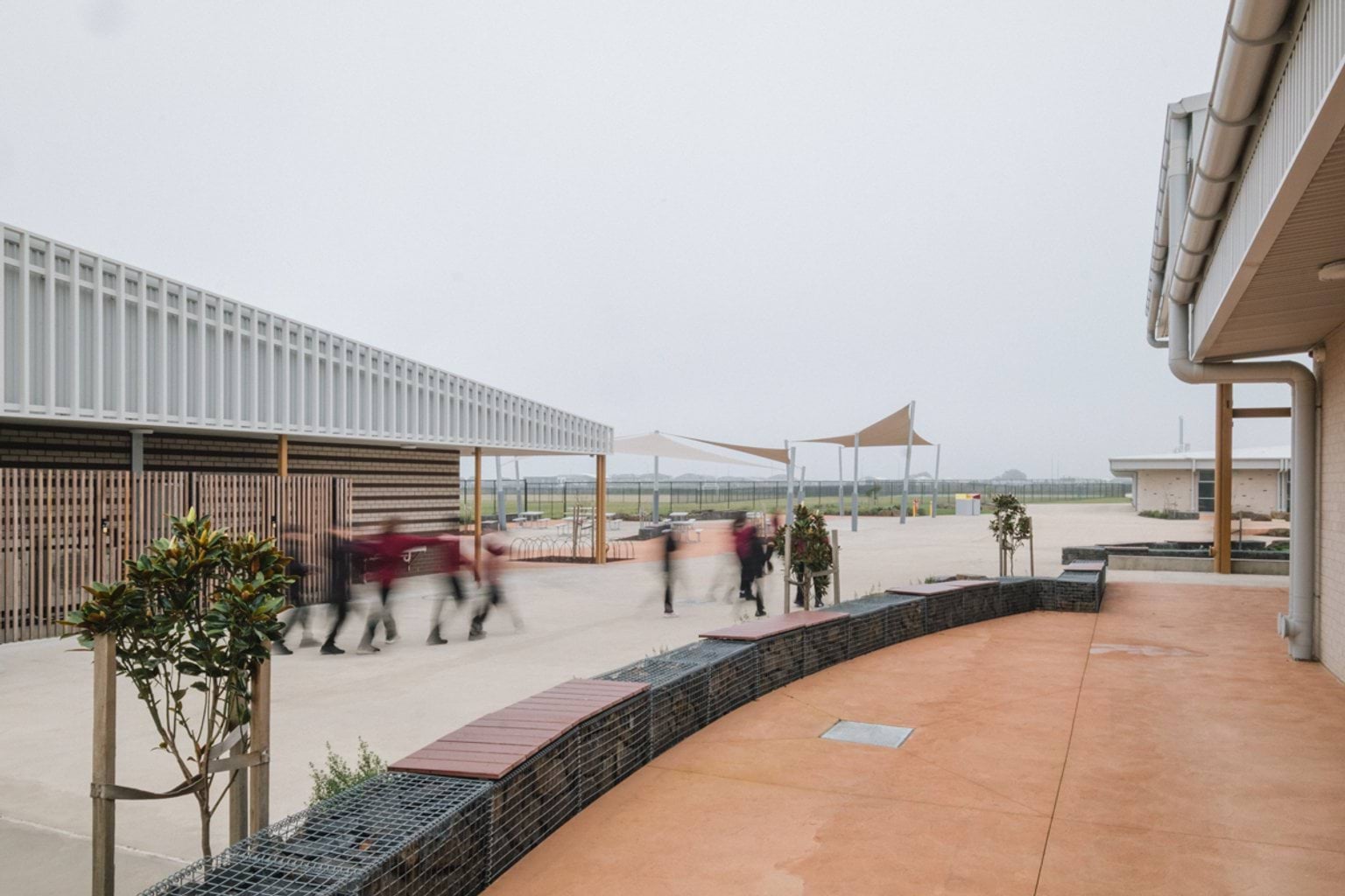Outdoor paved area between two buildings with curved benchtops, lined with trees. We can see students playing and sheltered play areas in the background