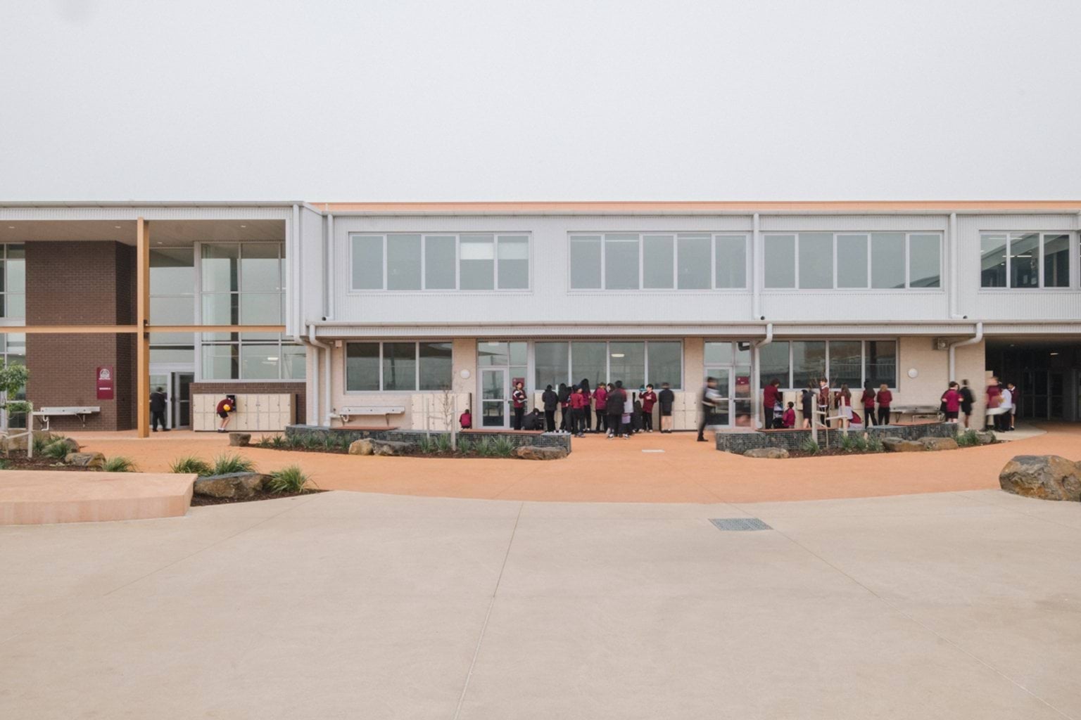 Outdoor paved area with school building in the background and bench tops and garden beds throughout. We can see students near the building entrance, accessing lockers.