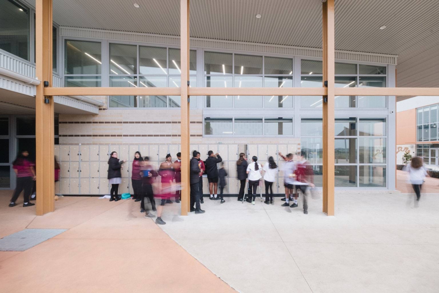 Outdoor covered area in front of building with orange beams, paved concrete and lockers. We can see students standing in open area