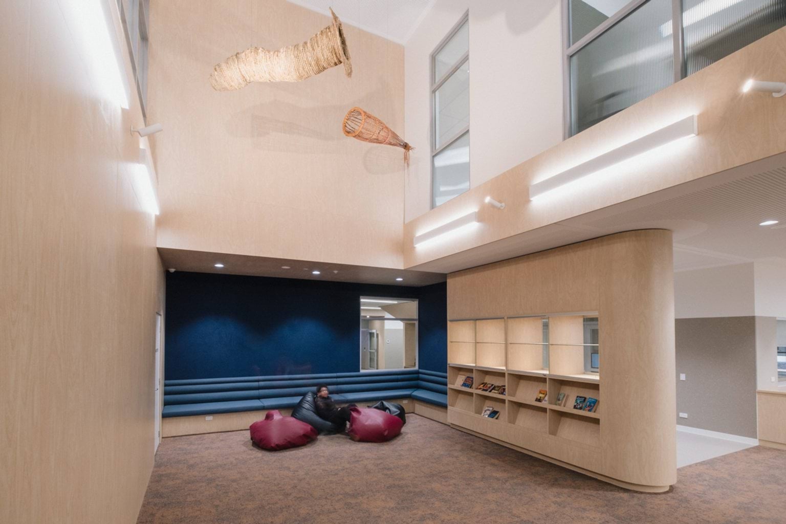 Open area with wooden curved walls, bookshelves, hanging art pieces and a blue feature wall. We can see beanbags with a student seated on one and carpet throughout