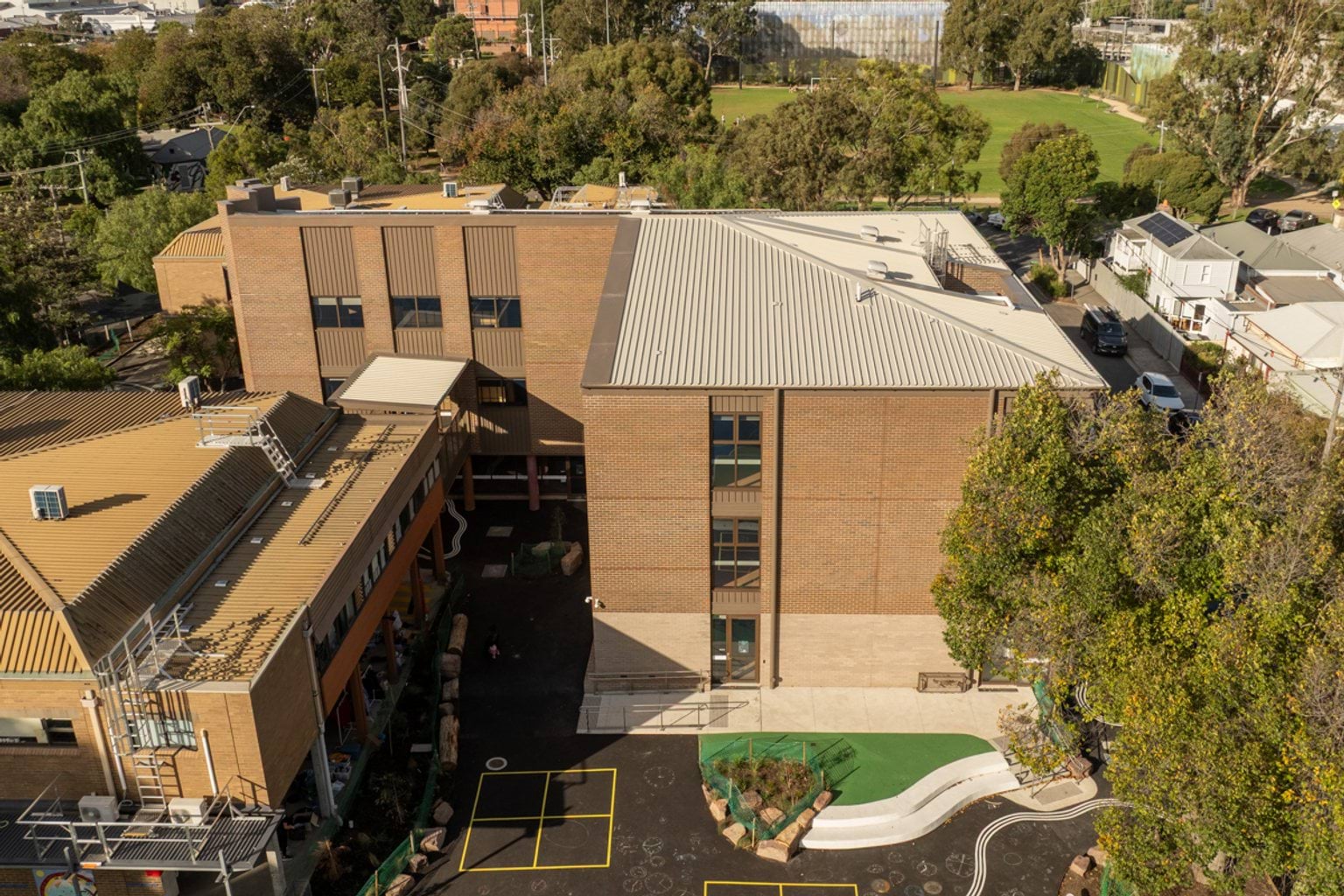 Birds eye view of two brown brick buildings with outdoor concrete play area surrounded by trees, lawn and neighbouring houses