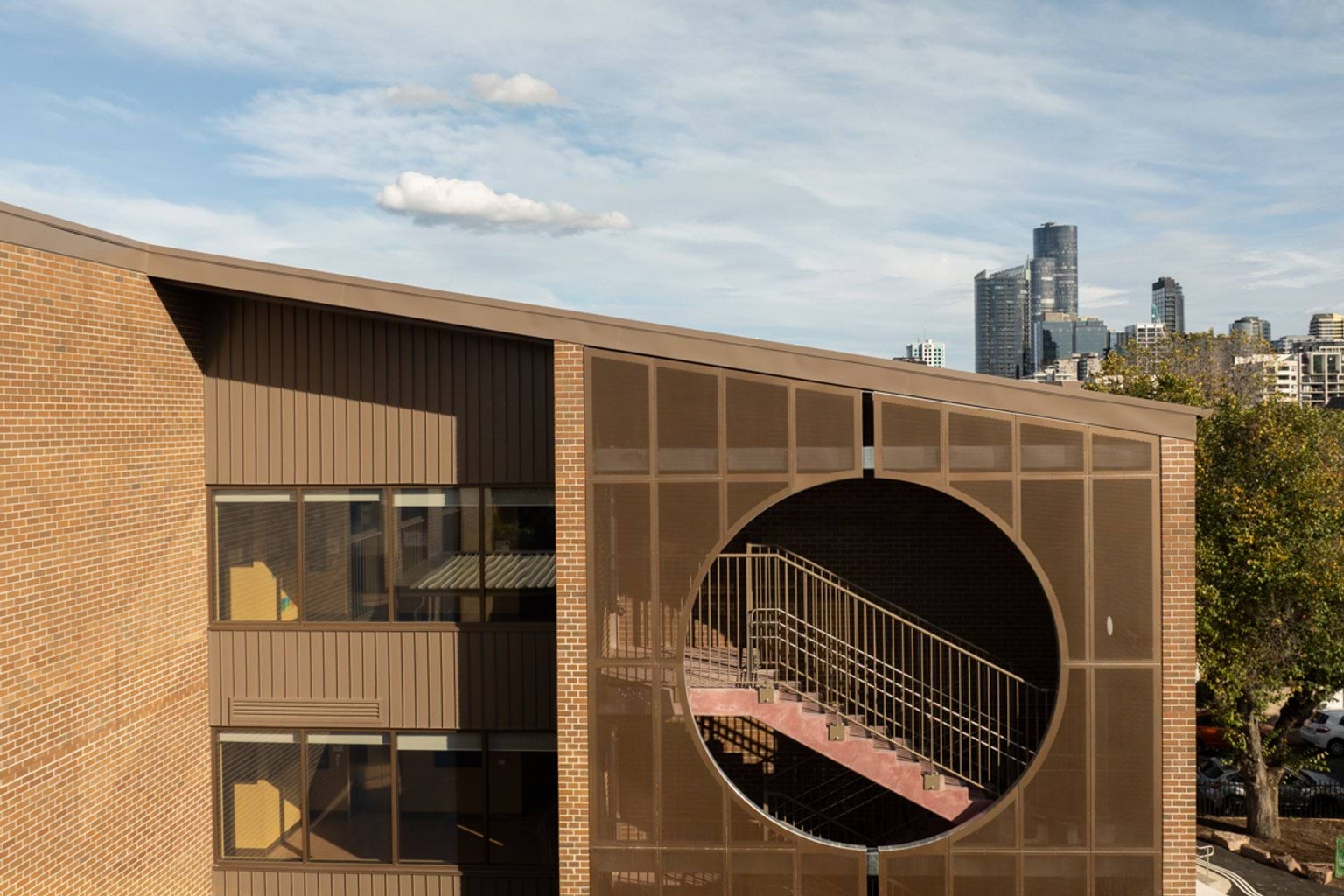 Brown brick building with slanted roof, windows and metal circular feature screen showing stairwell inside. We can see the partly cloudy city skyline and a tree in the background