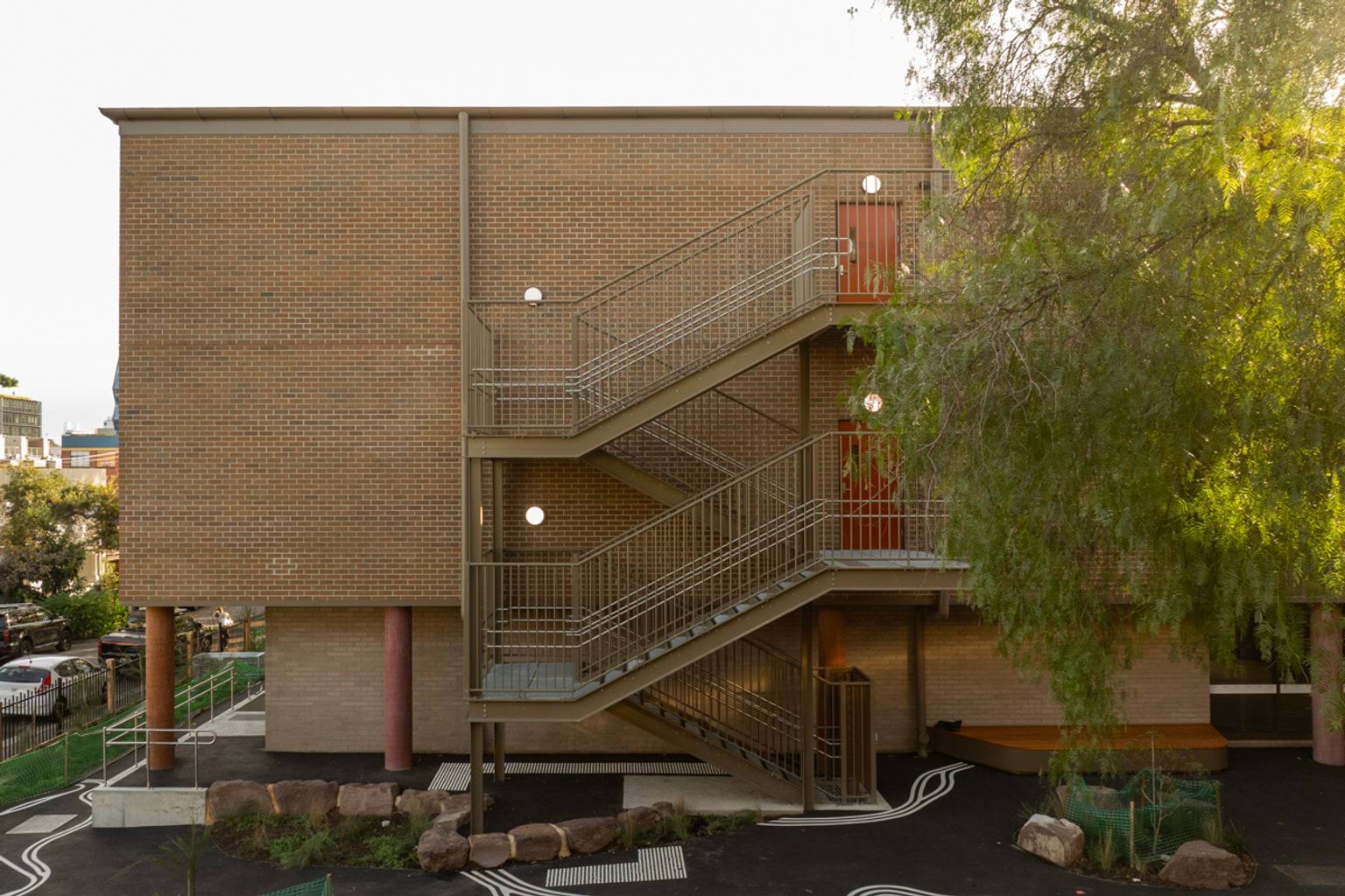 Side view of brown brick building with four flights of stairs on the outside. Large tree on the right side and concrete paved area below