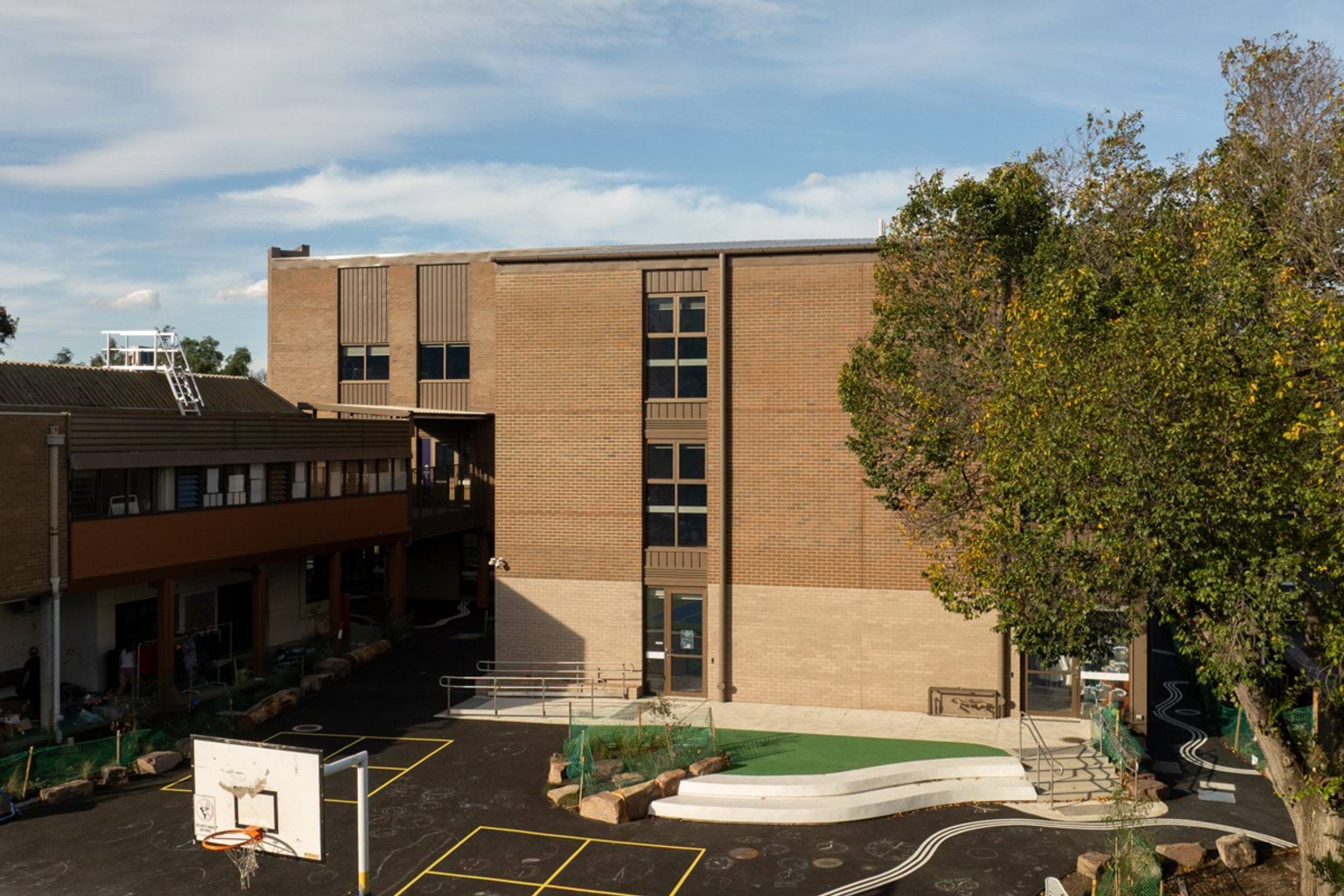 Side view of brown brick building with large tree and basketball ring in the foreground, steps and ramp on paved area
