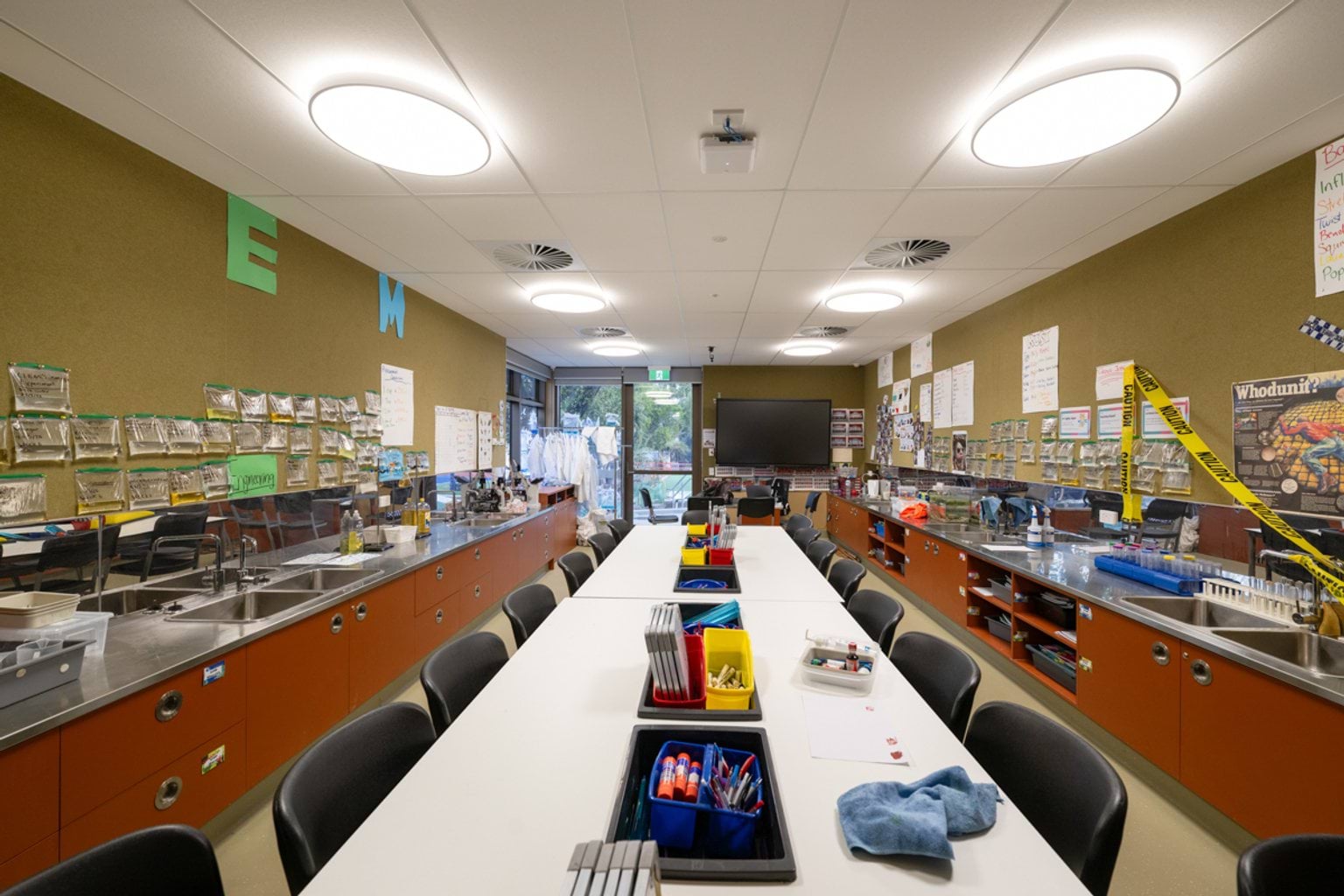Science classroom with long table in centre of classroom with chairs along both sides and benchtops and sinks on each side wall