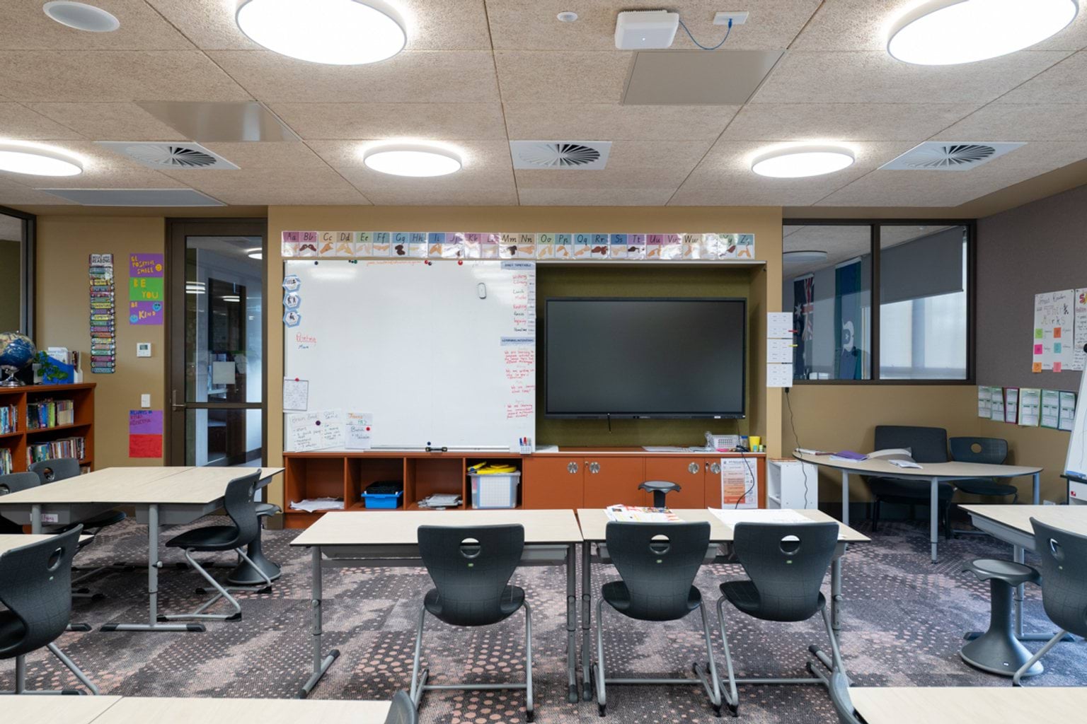 Classroom with grey patterned carpet, a screen and whiteboard with orange cabinets below and tables and chairs throughout
