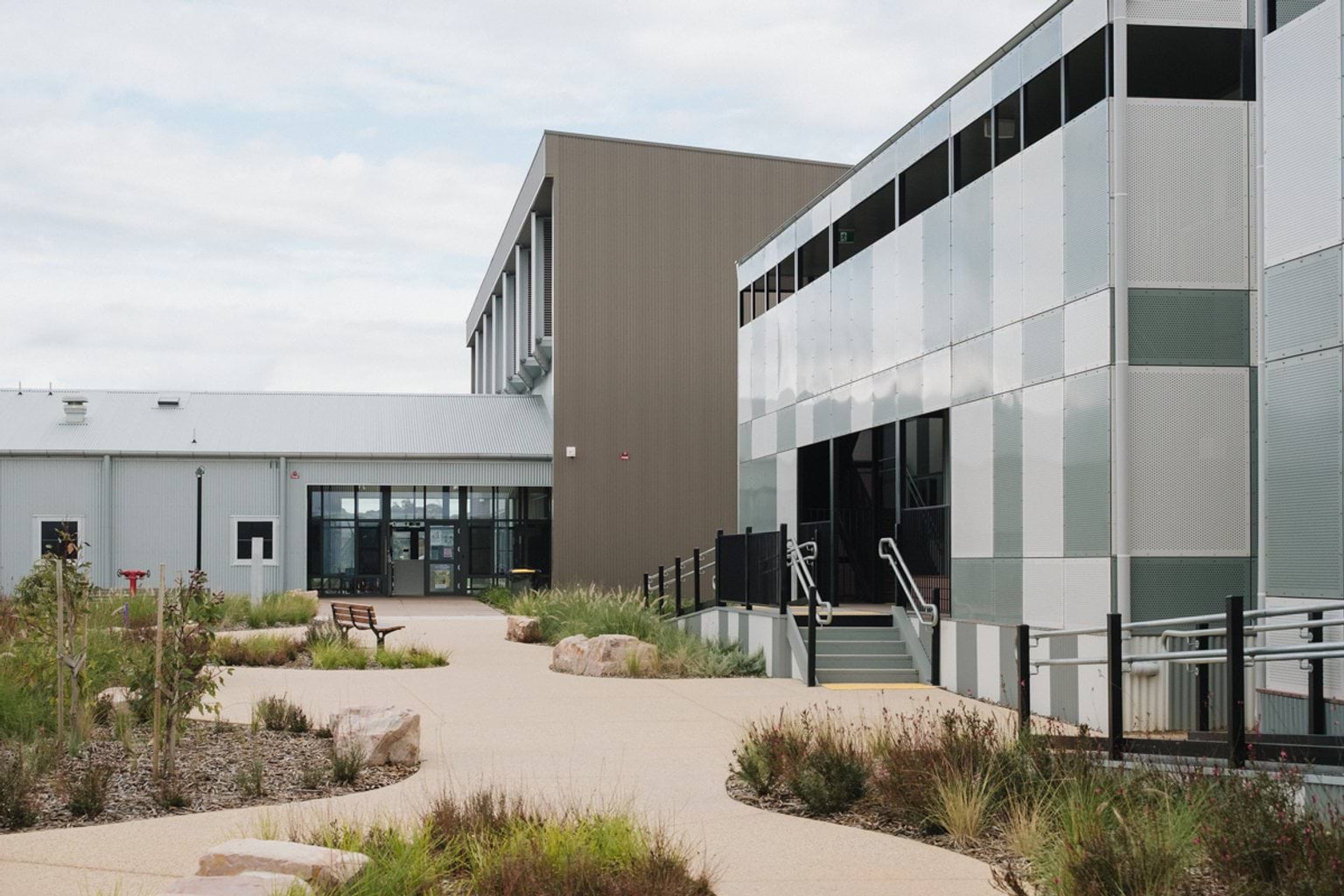 Outdoor paved area with garden beds and a bench seat. We can see stairs and ramps leading to two buildings with grey, green and brown panelling  