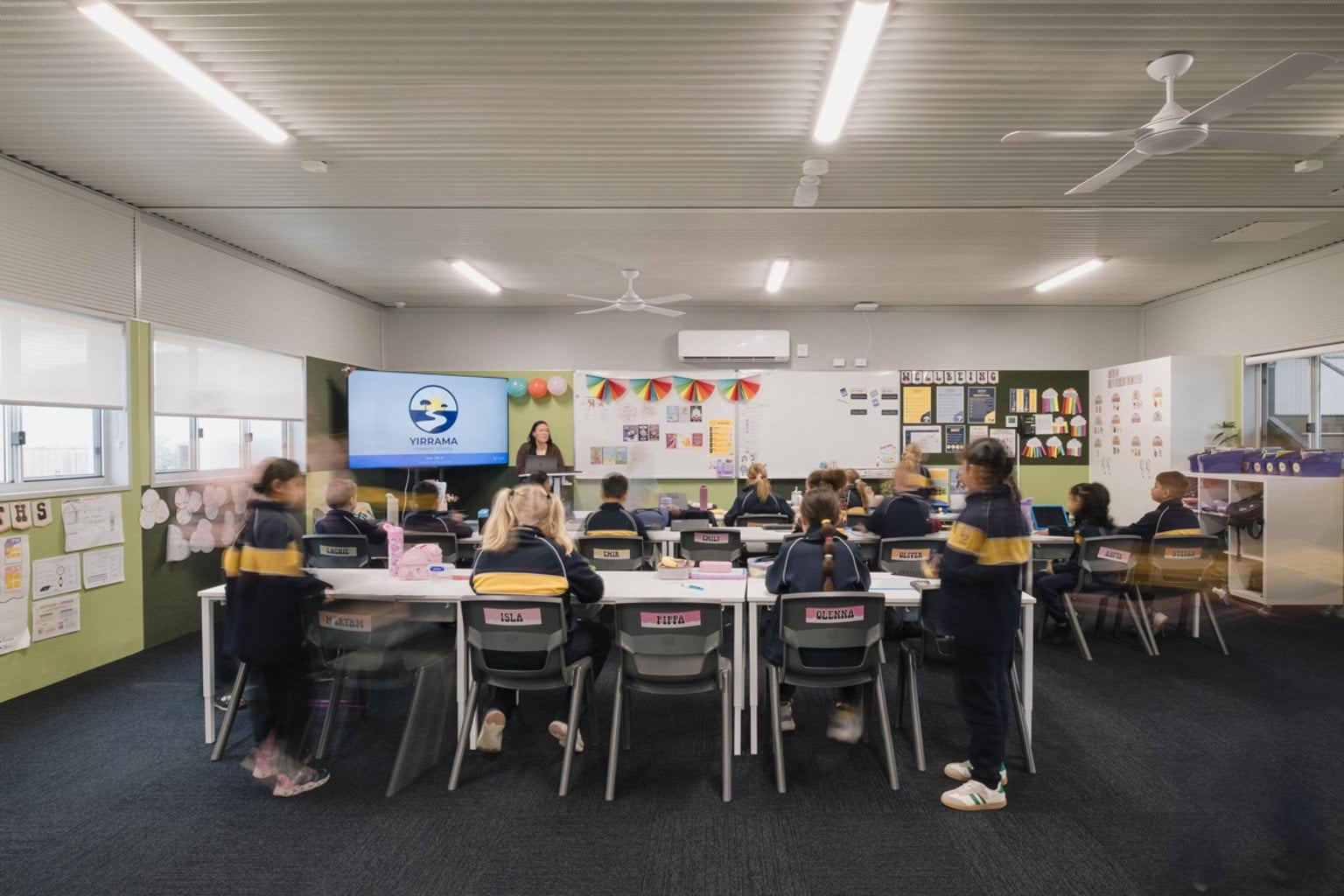 View of classroom from back of the room. We can see students seated along rows of tables and chairs, dark blue carpet and light green walls. There's a screen and a whiteboard at the front of the room