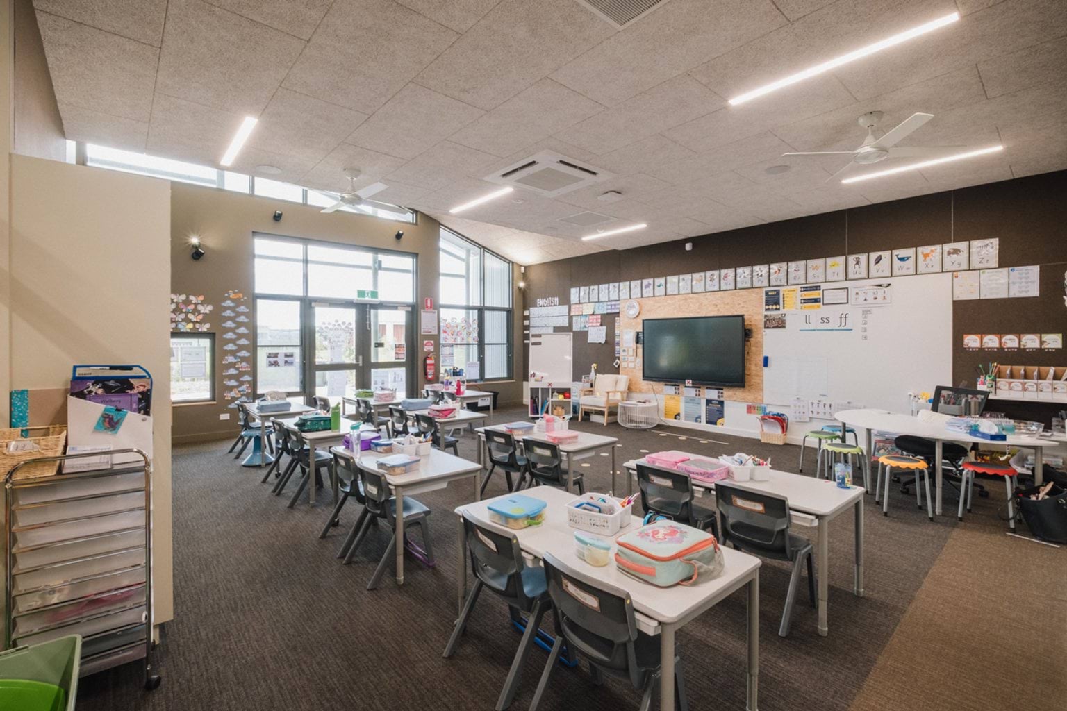 View from corner of classroom with brown carpet and brown walls. We can see rows of tables and chairs with lunchboxes. At the front of the room is a whiteboard and screen.