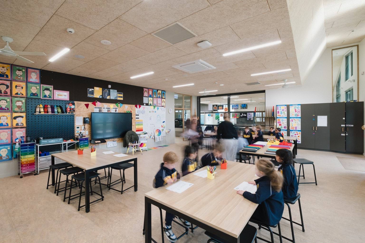 Art classroom with tables and chairs throughout. We can see students seated with pieces of paper. Cream coloured floor and a screen and whiteboard at the front of the room