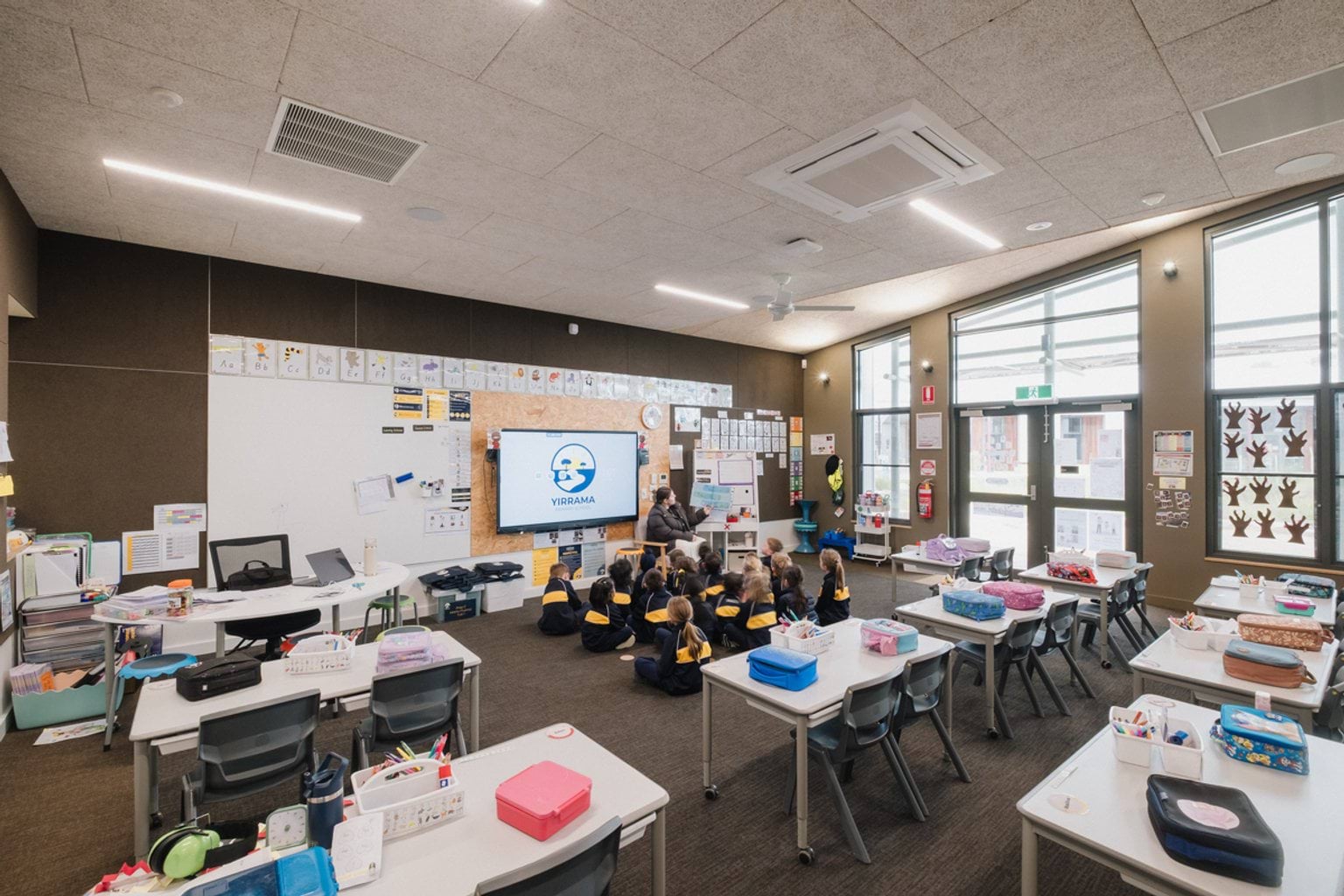 Classroom with brown carpet and brown panelling on the walls. We can see rows of tables and chairs and students seated on the floor at the front of the classroom.