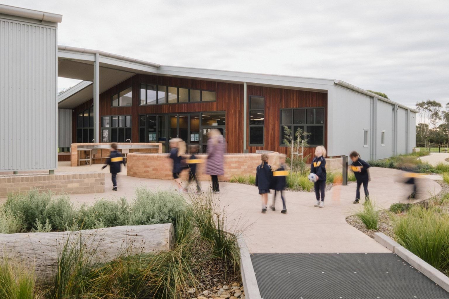 Students and a teacher walking along footpath outside school building. We can see garden beds surrounding the path