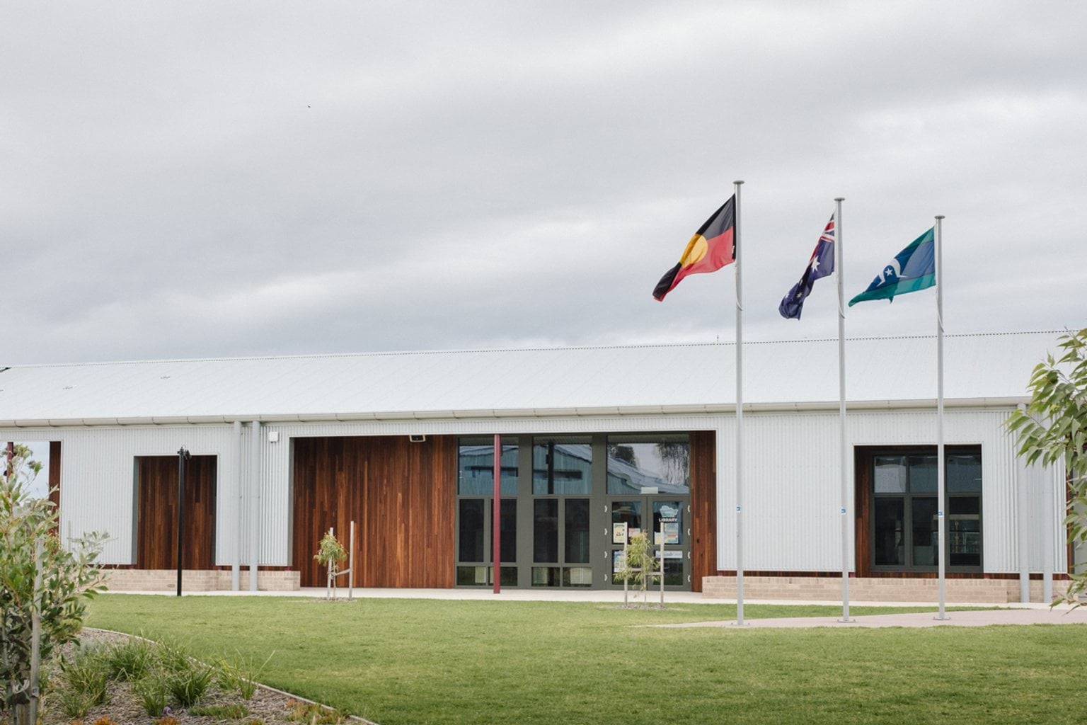 Single storey white building with wooden panelling and windows. In the foreground we can see Indigenous, a Torres-Strait and Australian flagpoles and grass lawn with garden beds