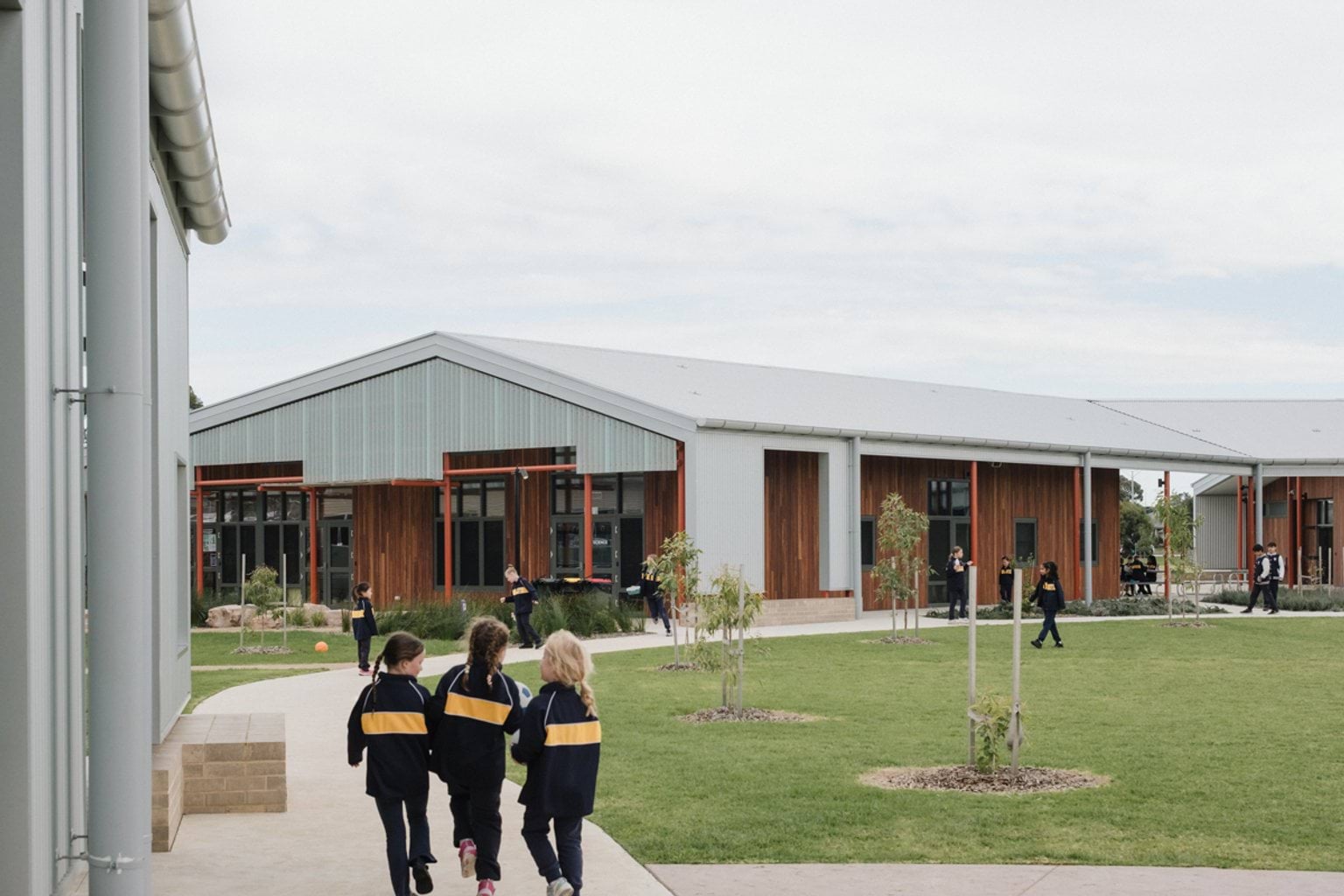 3 young students in uniform walking along paved footpath next to school building and grass lawn. We can see other students and another school building in the distance
