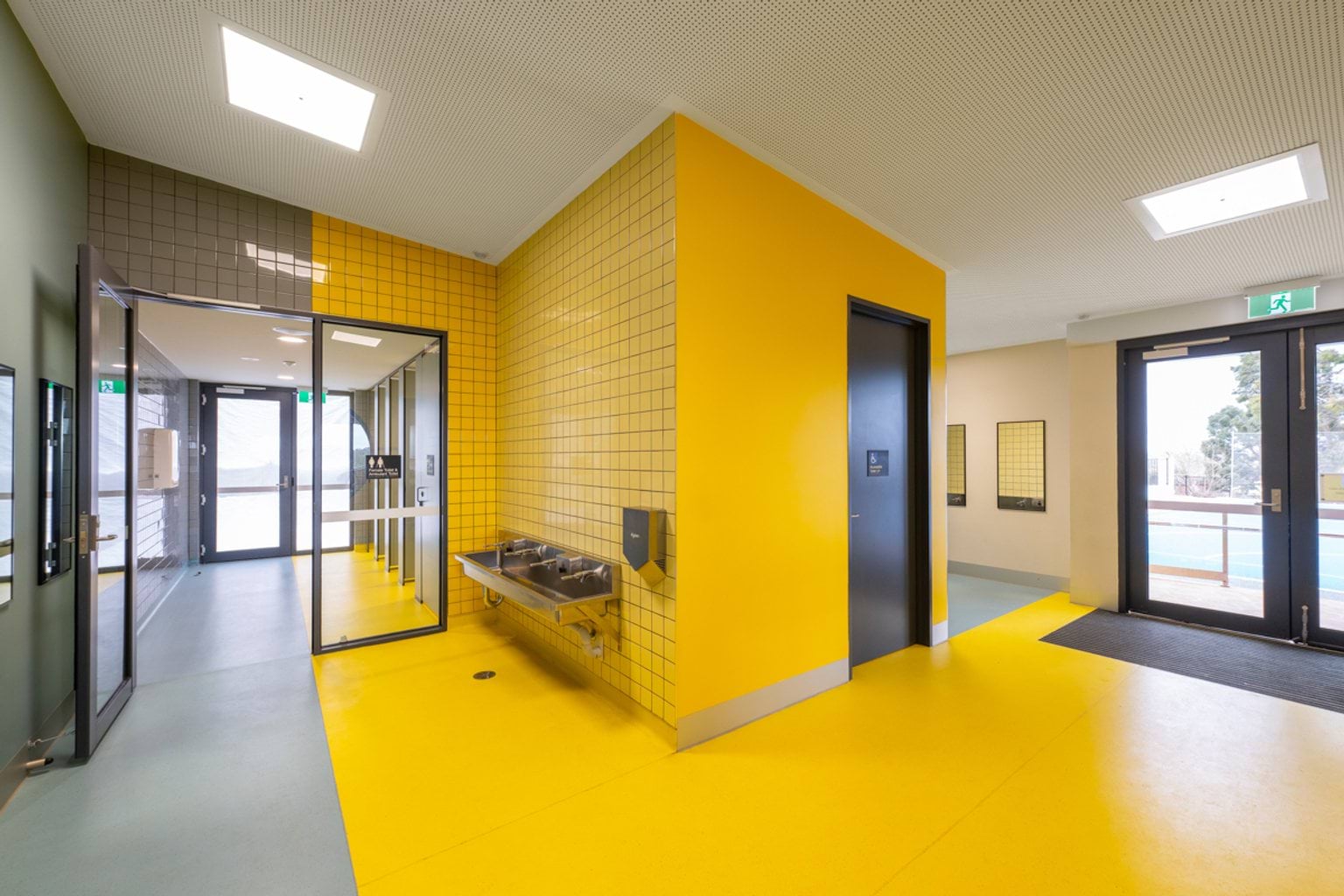 A hallway leads from an open glass door around a corner. There is a long sink with drinking fountains in the corner - this section is bright yellow.