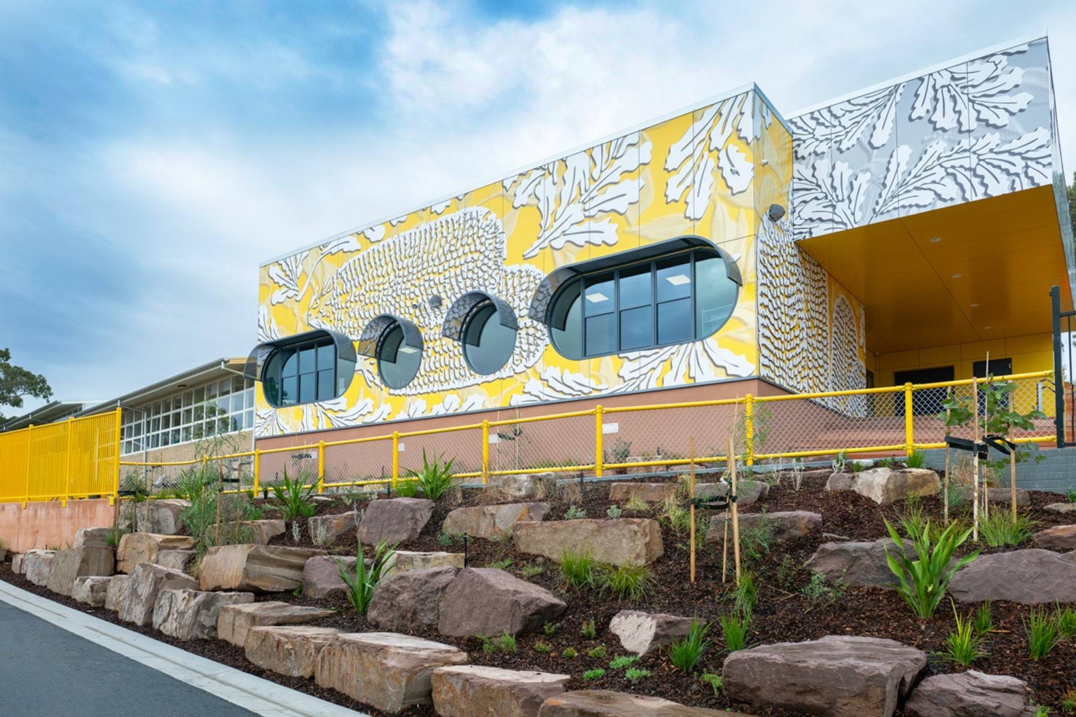 A large yellow building covered in white images of banksia flowers and leaves. In front of the building is a landscaped area with rocks and plants.
