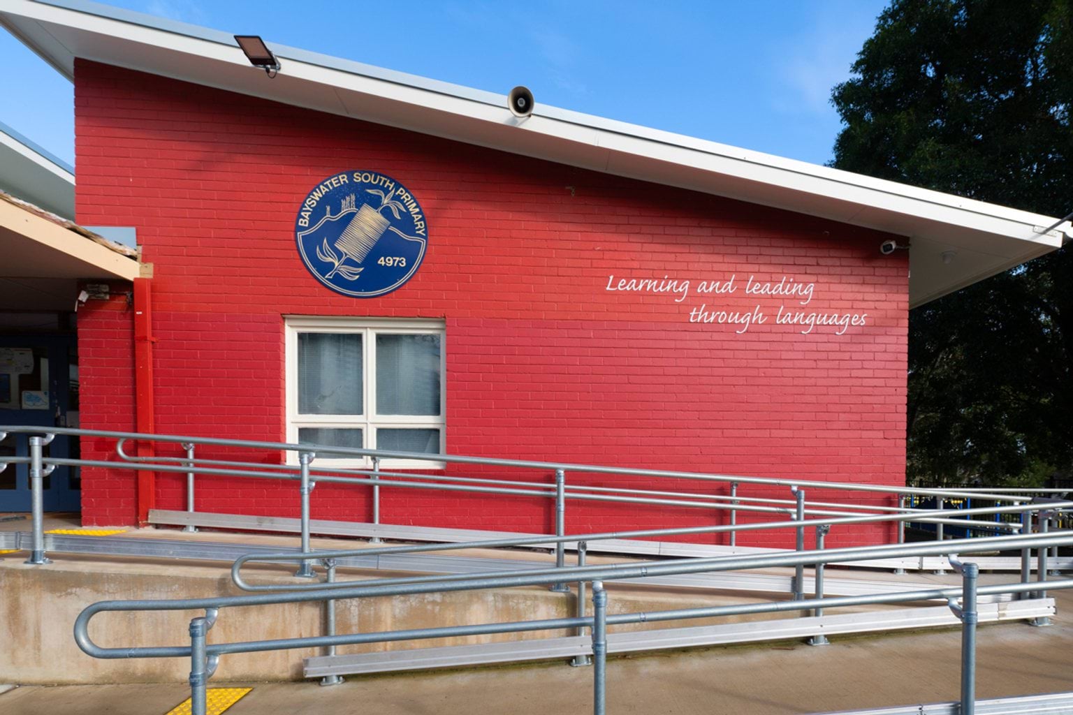 A ramp with metal railings leads up to a red, asymmetrical building. The building features Bayswater Primary School's blue and yellow logo. We can also see their motto: "learning and leading through language" on the wall.