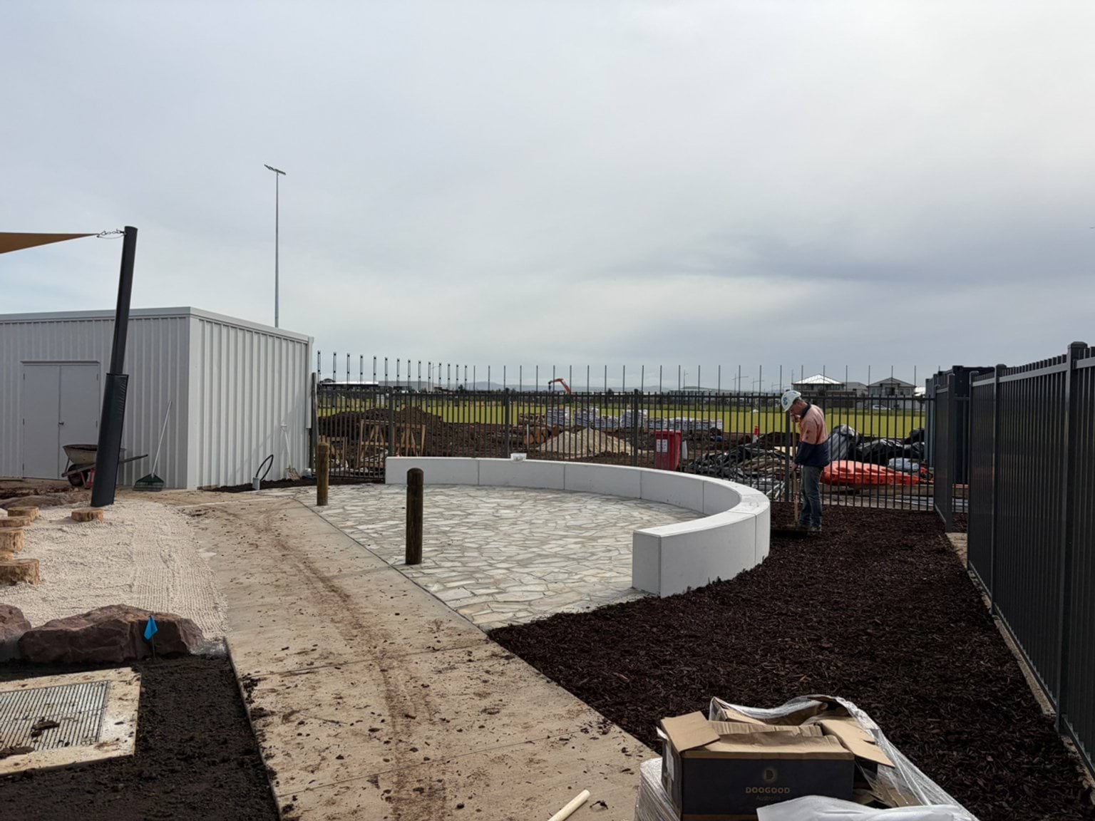 An outdoor landscaped area under construction featuring paved paths, garden beds, and surrounding fencing.