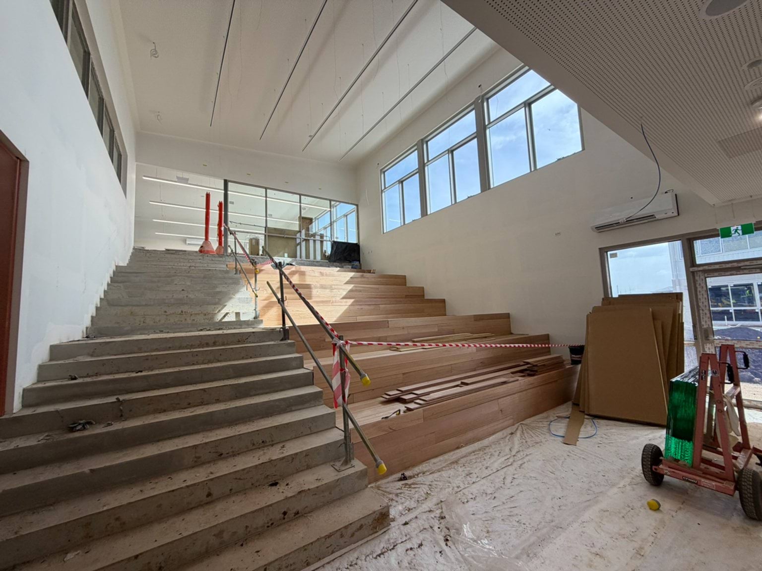 A bright, unfinished interior staircase with timber seating steps, handrails, large windows, and construction materials visible throughout the space.