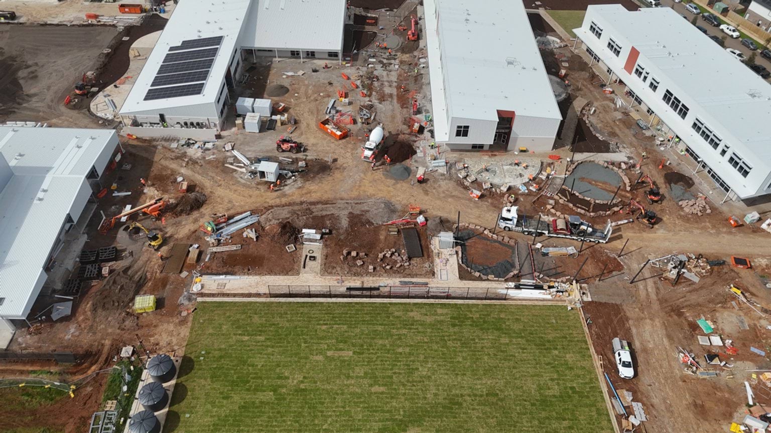 An aerial view of a construction site showing several large buildings, active earthworks, construction materials and a sports field in the foreground.