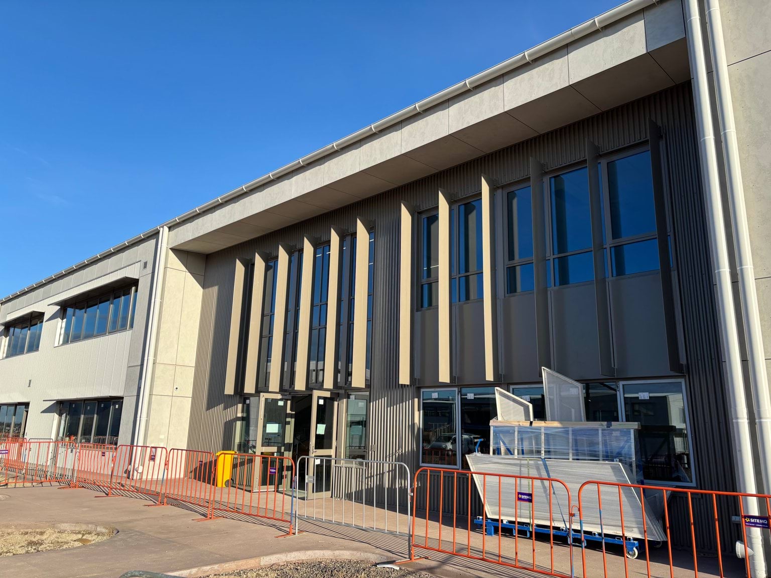 A building entrance under construction with tall windows and temporary fencing along a new paved walkway