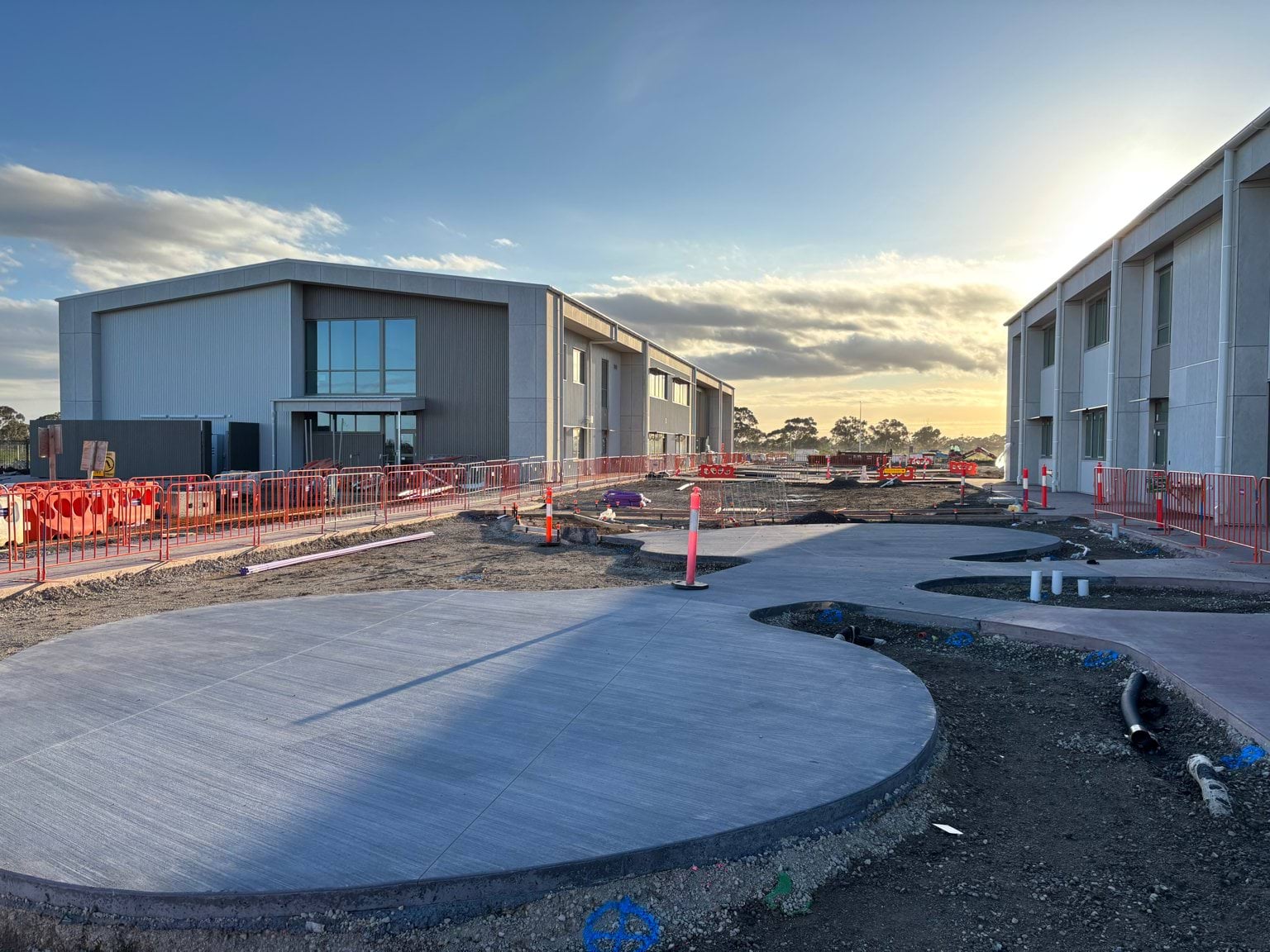 A curved concrete pathway between grey buildings with construction fencing and unfinished landscaping visible.