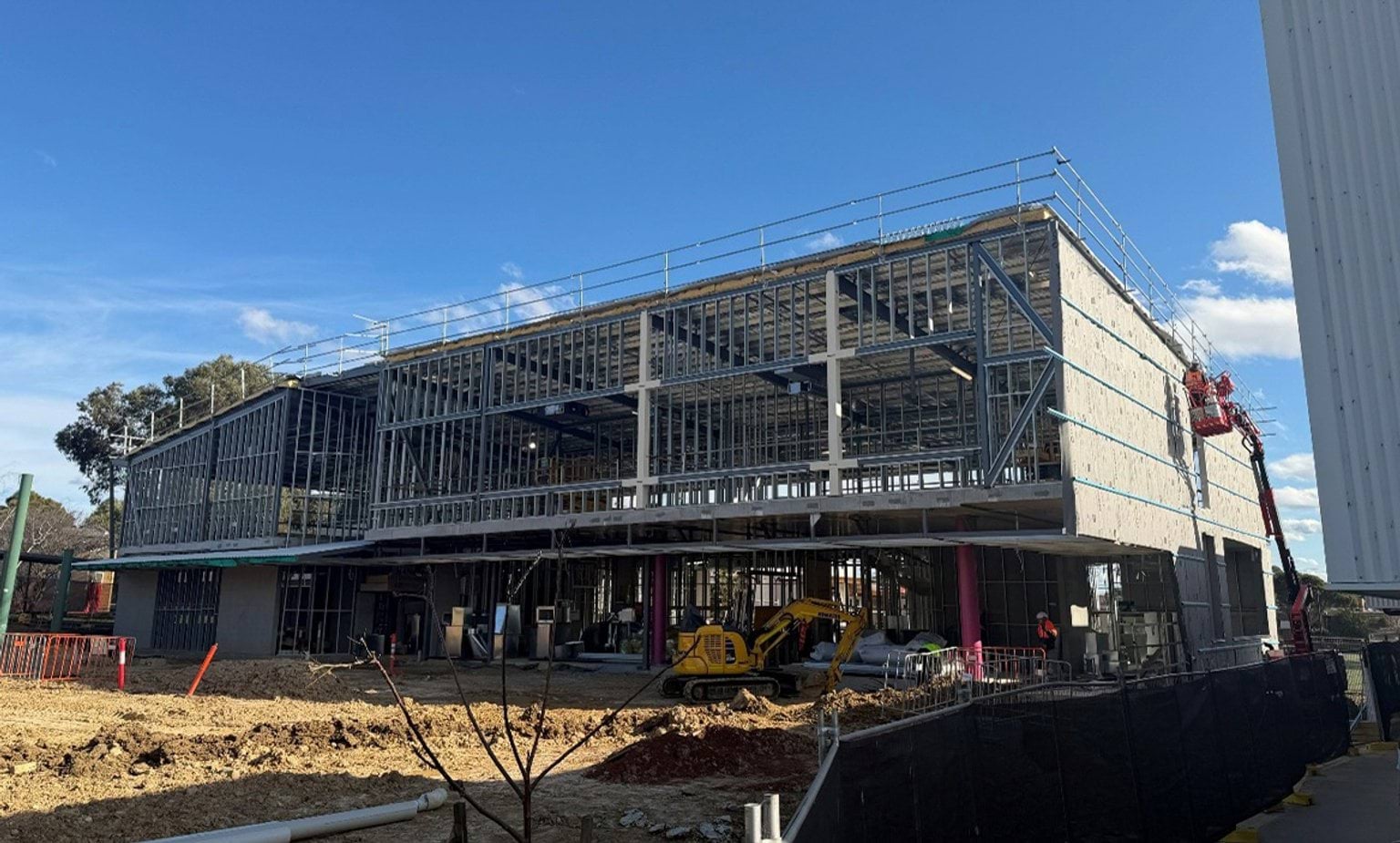 Ground-level view of a two-storey building under construction, showing the exposed steel frame, scaffolding and construction equipment on a worksite.