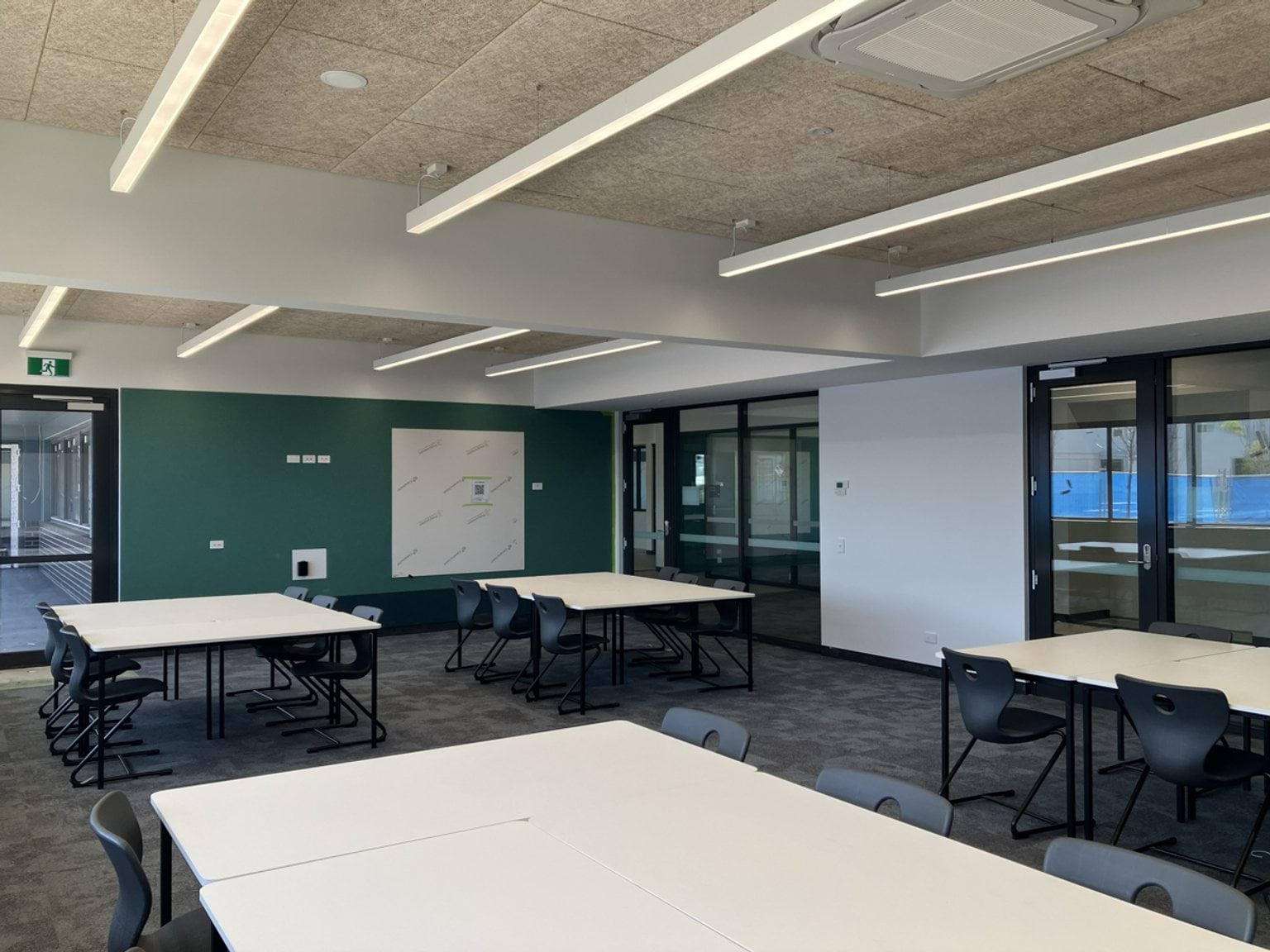 A classroom with tables and chairs. The room includes large windows and overhead lighting.