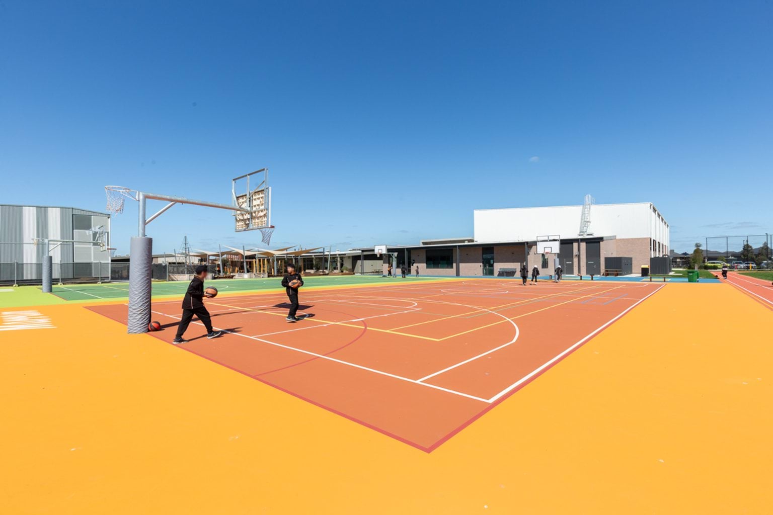 Multi-coloured outdoor court areas with basketball and netball rings. We can see a school building in the background and two students playing on the court.