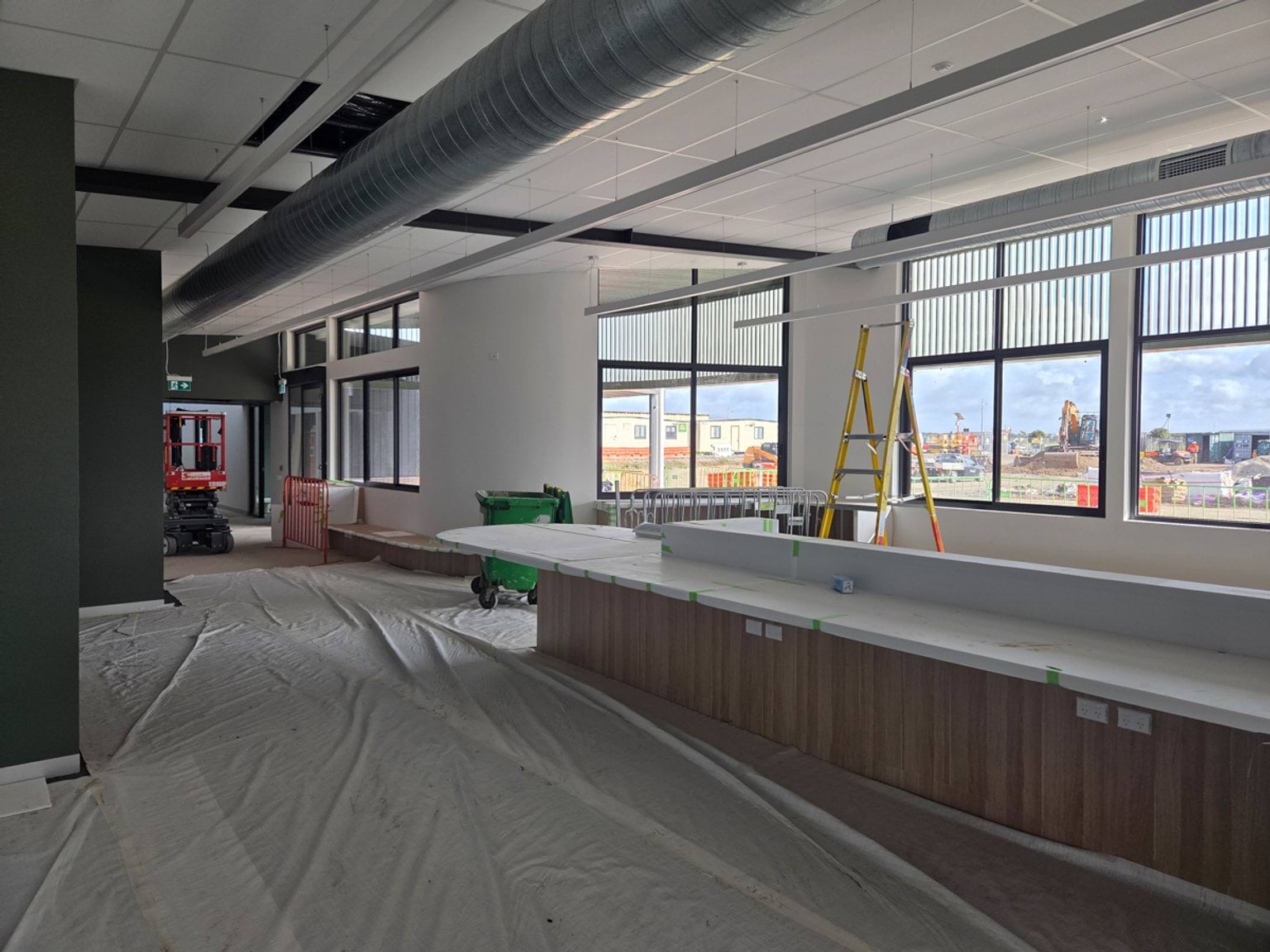 Foyer area under construction featuring a long built-in bench or counter beneath large windows. Protective floor coverings, a ladder, exposed ceiling services and unfinished ceiling panels and large windows are visible.