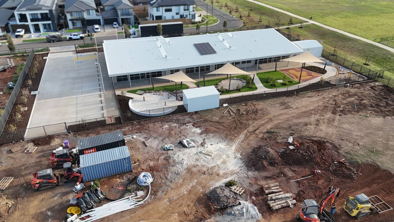 Aerial view of a school building under construction, with completed buildings surrounded by exposed soil, construction materials and earthworks.